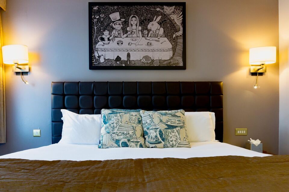bedroom at mad hatter hotel, double bed with lamp either side and a painting of alice in wonderland above the bed
