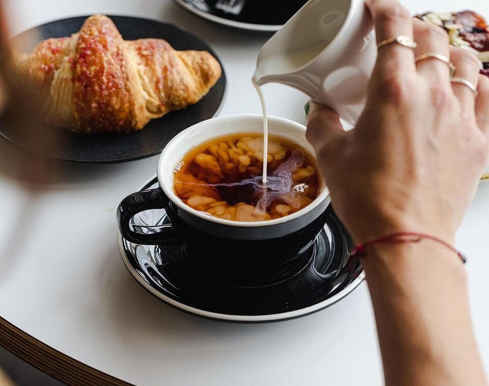 a close up shot of a hand pouring a small jug of milk into a cup of black coffee on a table with a croissant in the background