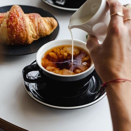 a close up shot of a hand pouring a small jug of milk into a cup of black coffee on a table with a croissant in the background