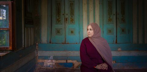 Muslim woman wearing a light pink long hijab sits in a room lined with blue painted wooden shutters and looks to her left out of an open window