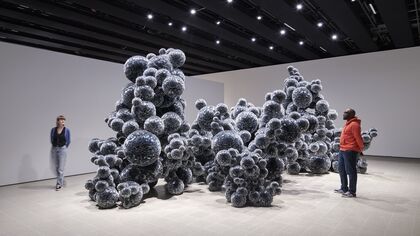 Large installation in gallery, silver grey ball like forms come together in an undulating bubbly mass. Two gallery visitors look on