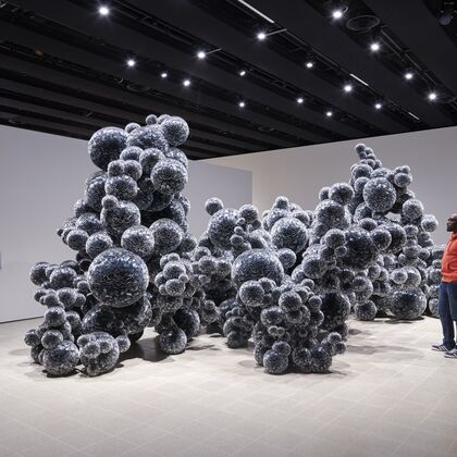 Large installation in gallery, silver grey ball like forms come together in an undulating bubbly mass. Two gallery visitors look on