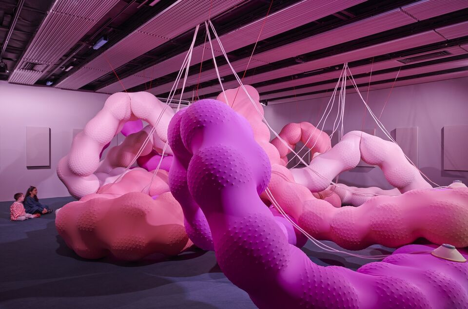 Huge bublous snake-like forms made from colourful pink rubber like material consume the central space of a large gallery. two children sit on the floor and look on