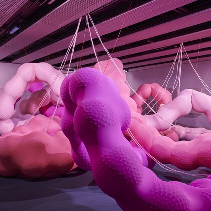Huge bublous snake-like forms made from colourful pink rubber like material consume the central space of a large gallery. two children sit on the floor and look on