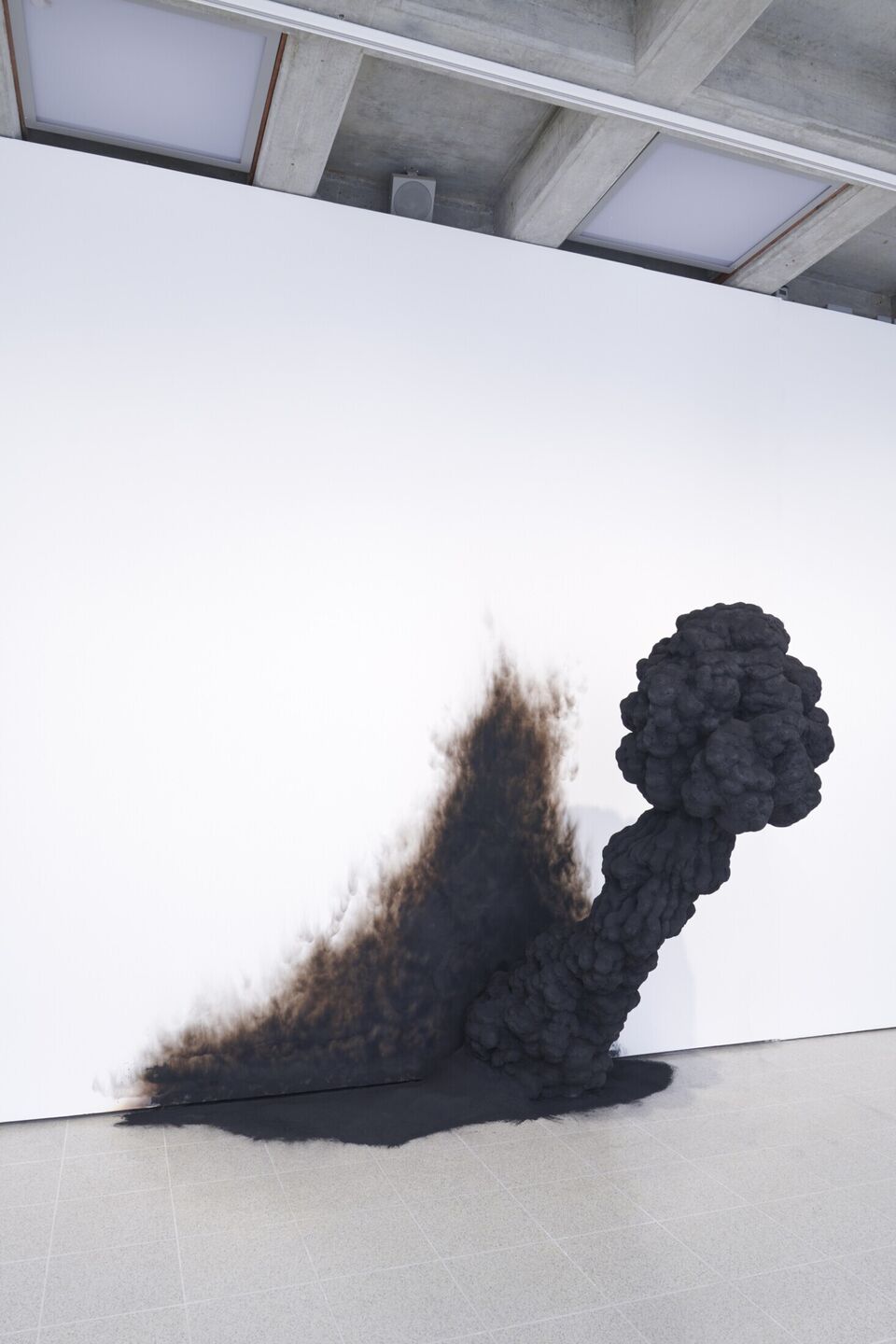 A charcoal black smokelike figure appears to emerge through ashes through the white wall of an art gallery