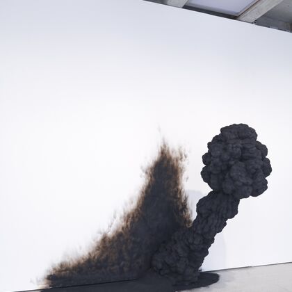 A charcoal black smokelike figure appears to emerge through ashes through the white wall of an art gallery
