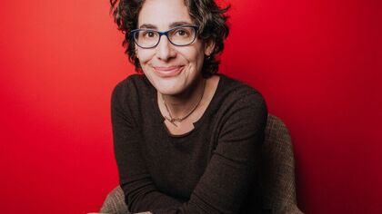 Sarah Koenig sits in front of a red background wearing a grey top