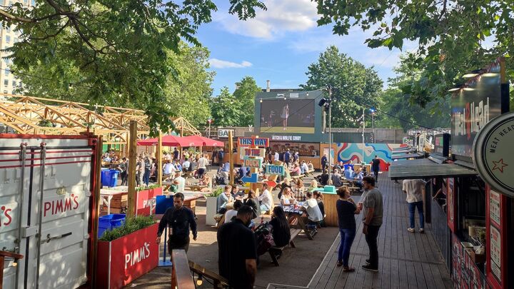 Ciew of people sitting in a sunny beer garden that is surrounded by trees. There is a large TV wall at the end of the beer garden showing sports