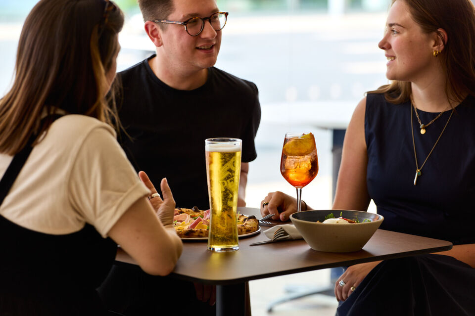 3 people sit at a high table with a glass of beer, a cocktail and a pizza