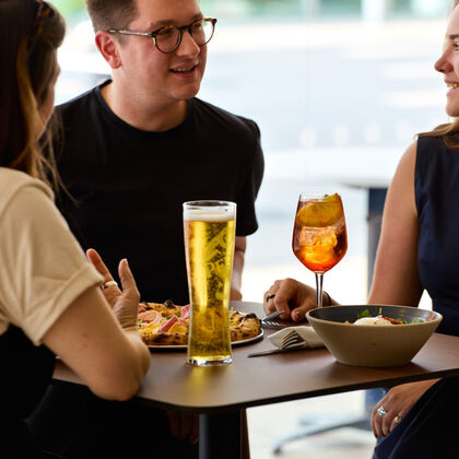 3 people sit at a high table with a glass of beer, a cocktail and a pizza