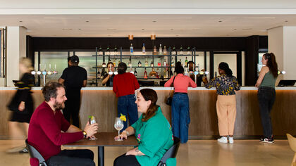 2 people sit at a table with drinks in the foreground, in the background people wait to buy drinks at the bar