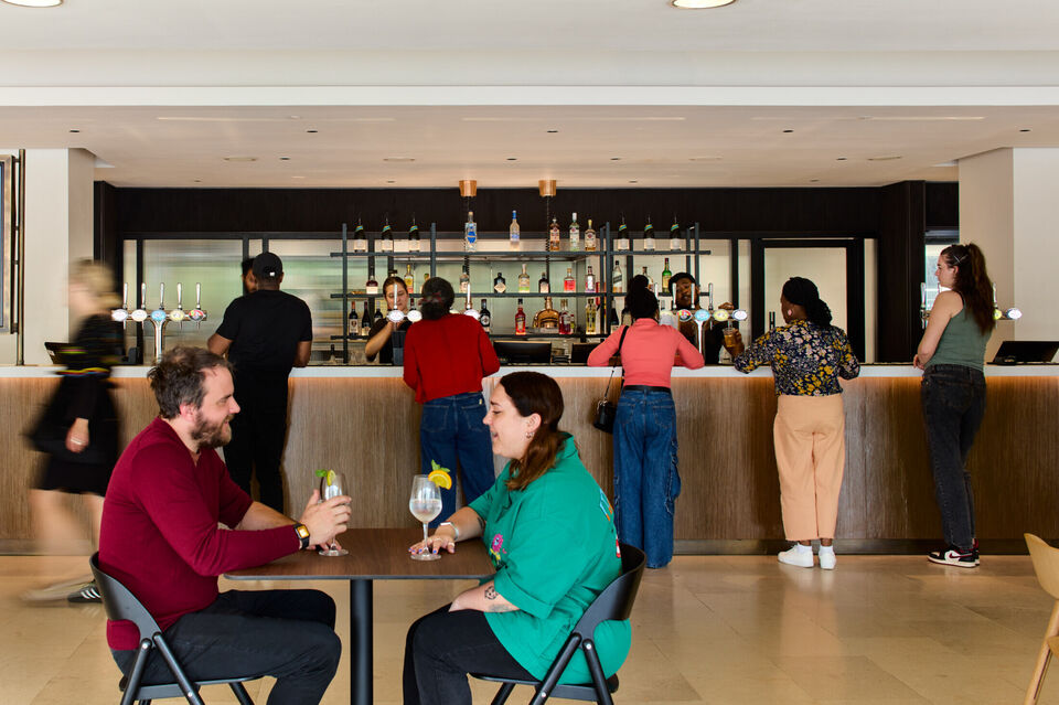 2 people sit at a table with drinks in the foreground, in the background people wait to buy drinks at the bar