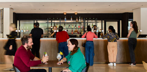 2 people sit at a table with drinks in the foreground, in the background people wait to buy drinks at the bar