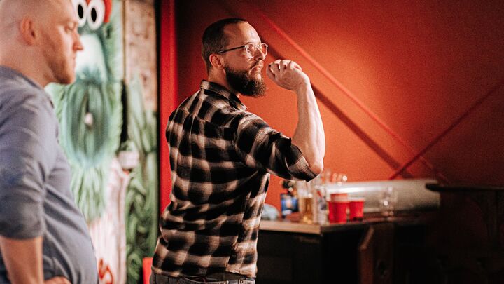 A side view a bearded man throwing darts, in the background pints of beer stand on a bar table