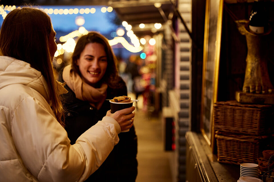 Two women buy a mulled wine from a market stall at night