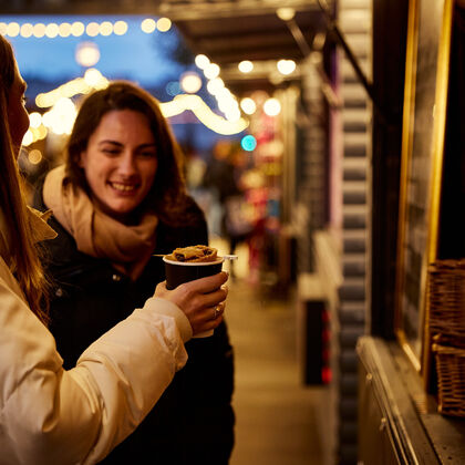 Two women buy a mulled wine from a market stall at night