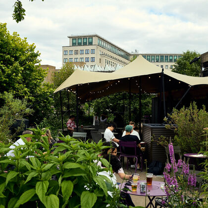 People sat drinking at an outdoor bar with a shelter, surrounded by plants and flowers.