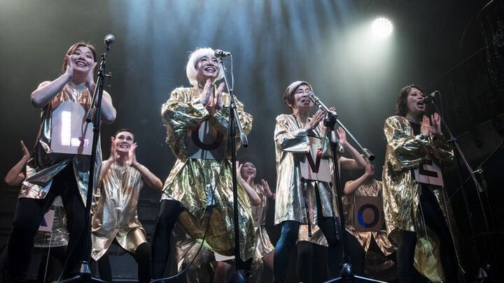 Performers on stage wearing gold and a letter on their outfit smiling and singing into microphones, the letters spell 'love