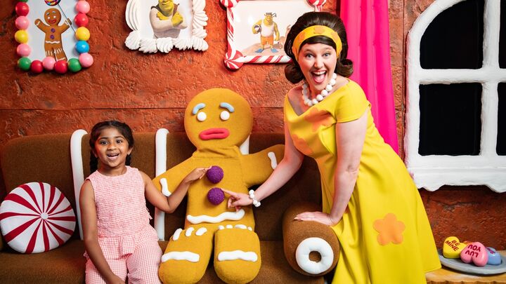 A little girl, a human-sized gingerbread man and a smiling woman wearing a yellow dress sit inside a gingerbread house decorated with sweets