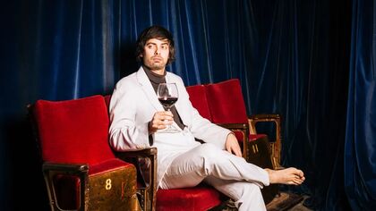 Pretentious man in white suit sitting on a red chair holding a glass of red wine