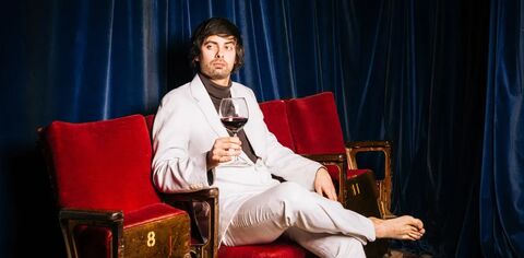 Pretentious man in white suit sitting on a red chair holding a glass of red wine