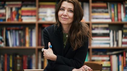 A woman with long hair wearing a black jacket and turquoise and silver jewellery - Elif Shafak - leans on a desk and looks at the camera, behind her are shelves full of books
