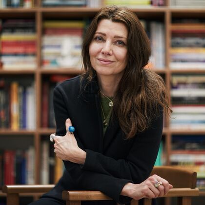 A woman with long hair wearing a black jacket and turquoise and silver jewellery - Elif Shafak - leans on a desk and looks at the camera, behind her are shelves full of books