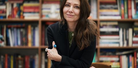 A woman with long hair wearing a black jacket and turquoise and silver jewellery - Elif Shafak - leans on a desk and looks at the camera, behind her are shelves full of books