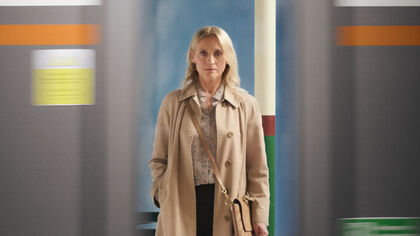 A woman stands wearing a trench coat and handbag and stares sternly through a train window
