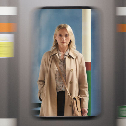A woman stands wearing a trench coat and handbag and stares sternly through a train window