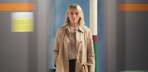 A woman stands wearing a trench coat and handbag and stares sternly through a train window