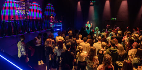 An audience watching a poet with a DJ off to one side, in a studio theatre with multi-coloured lighting