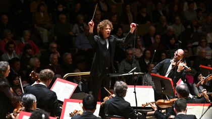 A conductor stands in front of an orchestra with hands raised the audience is visible in shadow in the background