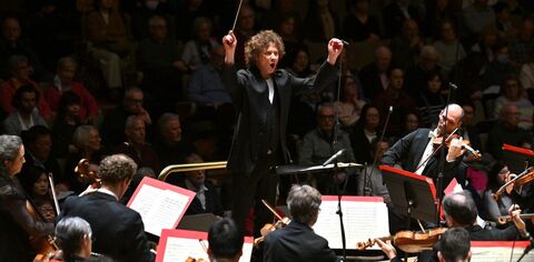 A conductor stands in front of an orchestra with hands raised the audience is visible in shadow in the background