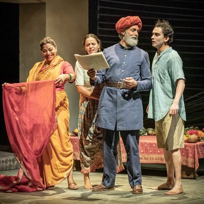 A bearded man in a red turban and a blue suit reads a paper to 2 women in saris and a young man in a green shirt on stage